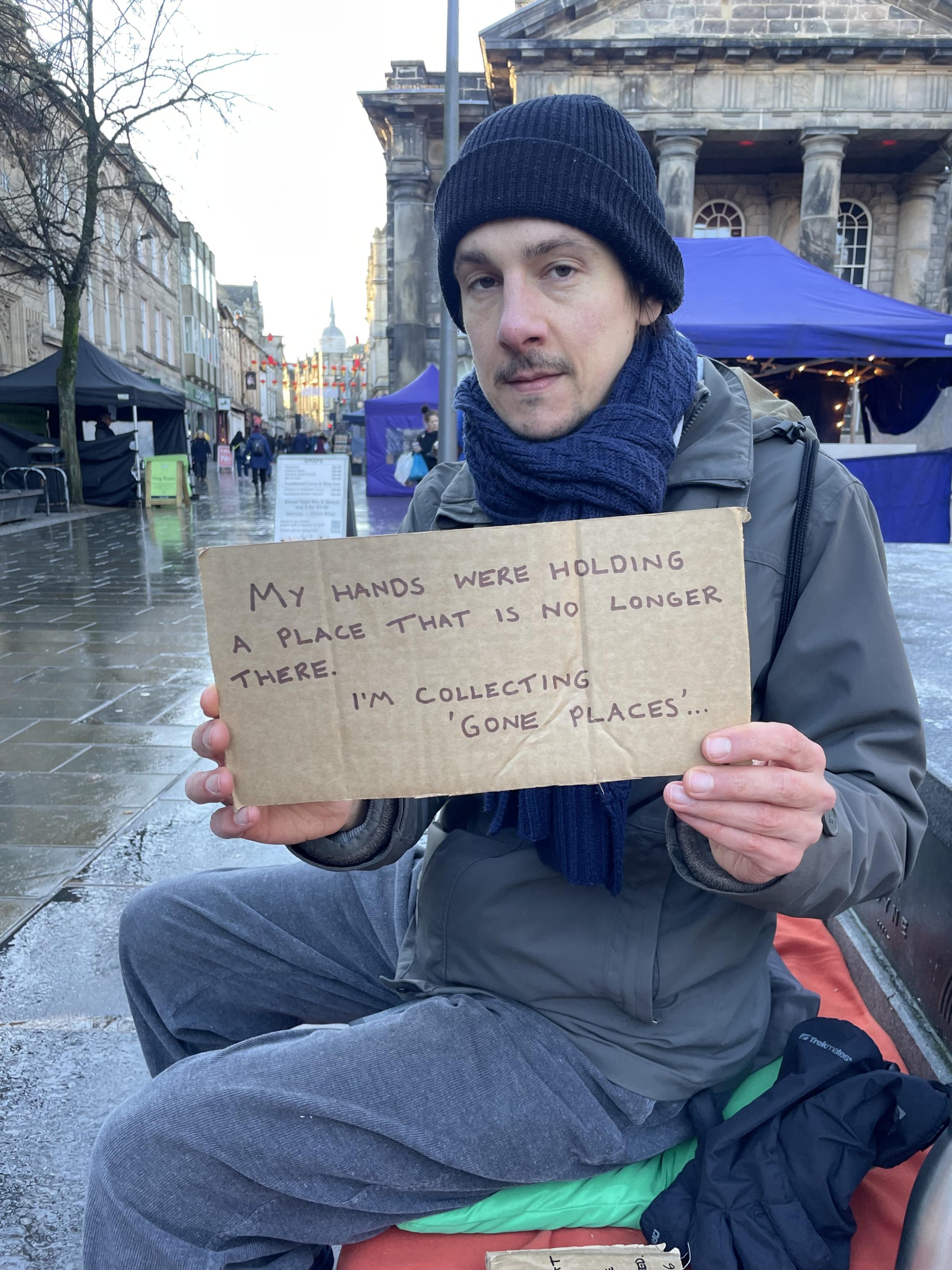 A man holding a cardboard sign that says "My hands were holding a place that is no longer there. I'm collecting 'Gone Places'.
