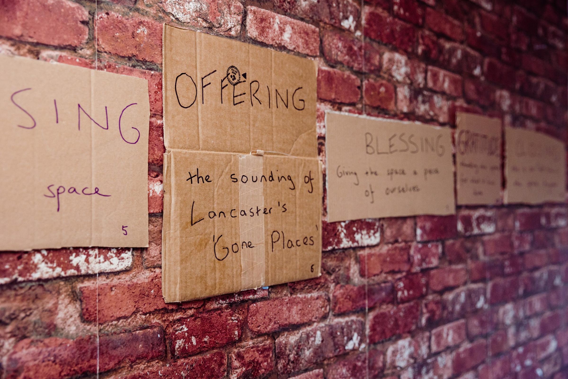 Cardboard signs on a wall that read "Sign Space. Offering. The sounding of Lancaster's Gone Places. Blessing. Giving the space a piece of ourselves"